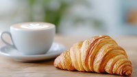 Fresh baked croissant on wooden table with cup of cappuccino coffee with latte art foam in white ceramic cup and saucer, morning breakfast scene.