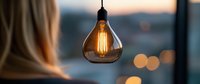 Glowing vintage Edison bulb with warm filament hanging against blurred evening cityscape background creating cozy atmospheric lighting and bokeh effect.