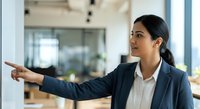 Young South Asian female professional in business attire pointing forward in modern office environment, suitable for corporate training, leadership, and business communication materials.