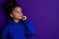 Young African American girl in blue hoodie looking up dreamily against purple background, hand on chin in contemplative pose, natural curly hair styled in bun.