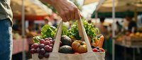 Shopper holding canvas tote bag filled with fresh organic produce including grapes, avocados, and vegetables at farmers market or grocery store.