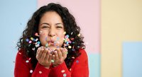 Young Hispanic woman in red sweater blowing colorful confetti from hands against pastel background for celebration and party concepts.