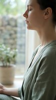 Young woman practicing mindfulness meditation by window with natural light and plants creating peaceful atmosphere for wellness and mental health.