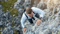 Young athletic male in business attire climbing steep rocky mountain face, combining professional dress with extreme sport activity, showing risk and challenge.
