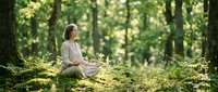Young woman practicing mindfulness meditation in peaceful forest setting surrounded by lush green ferns and tall trees creating serene natural wellness environment.
