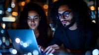 Two young African American professionals working together on laptop computer in modern office with warm bokeh lights creating cozy evening atmosphere.
