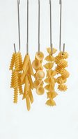 Assorted dried pasta shapes hanging from metal spoons against white background, showcasing various Italian pasta types for cooking and food styling.