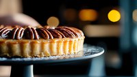 Chocolate pecan tart on dark plate with blurred bokeh background, close-up view showing flaky crust and glossy nutty topping in warm ambient lighting.
