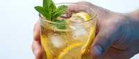 Hand holding refreshing iced tea glass with lemon slices and fresh mint garnish. Cold summer beverage for relaxation and hydration on white background.