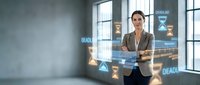 Young woman with dark hair and olive complexion, wearing a beige blazer, interacts with holographic digital interface displaying data analytics and deadline icons in modern office setting with large windows and city skyline view.