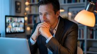 Middle-aged Caucasian businessman in suit working late at office desk with laptop and video conference call on screen in modern workspace with warm lighting.
