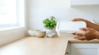 Hands holding white takeout container in bright kitchen with potted plant and window light, suitable for food delivery, restaurant, and catering marketing materials.