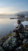 Contemplative man sitting on mossy rocks by misty lake at dawn with wooden cabin and boat in serene wilderness landscape for meditation and solitude concepts.