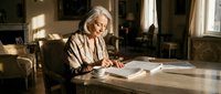 Senior Caucasian woman with gray hair and glasses reading documents at wooden table in sunlit home office, focused on paperwork and financial planning.