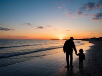 Adult and child silhouettes stand together on sandy beach at sunset, watching golden sun descend over ocean horizon with colorful sky. Family bonding moment.