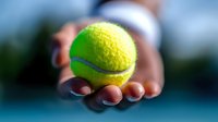 Bright yellow-green tennis ball held in outstretched palm against blurred blue court background, showcasing sports equipment for tennis match or practice.