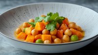 Chickpeas and butternut squash cubes in spicy sauce garnished with fresh cilantro leaves served in white ceramic bowl, healthy vegan meal on dark background.