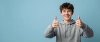 Happy teenage boy giving thumbs up gesture wearing gray hoodie on blue background expressing positive emotions and success.