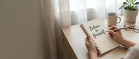 Woman writing believe in yourself motivational quote in notebook at wooden desk with coffee cup and plant creating inspiring workspace for personal development.
