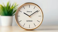 Minimalist wall clock with motivational text showing time to achieve on white dial, gold frame against blurred plant background. For office decor and productivity concept.