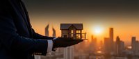 Businessman holding wooden house model against sunset city skyline backdrop representing real estate investment and property development opportunities.
