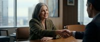 Professional mature businesswoman shaking hands with colleague in modern office meeting room during successful negotiation or partnership agreement.