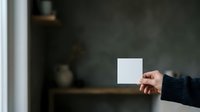 Person in dark sweater holding blank white card against blurred interior background, suitable for mockups, advertising campaigns, and promotional materials.
