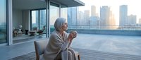 Mature woman in bathrobe enjoying morning coffee on modern apartment balcony with city skyline view. Relaxation and luxury lifestyle concept.