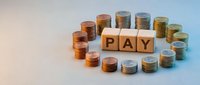 Stacked coins arranged in circle around wooden blocks spelling PAY on light background, representing payment, salary, wages, financial transactions and money management concepts.