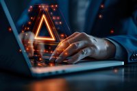Glowing warning triangle symbol with orange light emerging from laptop screen while person types, representing cybersecurity threat or digital alert in dark environment.