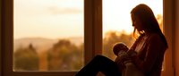 Young mother breastfeeding newborn baby by window during golden hour sunset creating intimate bonding moment in warm natural light for motherhood concepts.