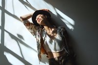 Young Asian woman in black hat and floral kimono enjoying sunlight through window blinds, creating dramatic shadow patterns on wall, expressing joy and serenity.