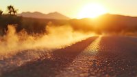 Dramatic sunset over empty mountain road with golden sunlight and dust particles creating atmospheric haze effect, close-up view of asphalt surface.