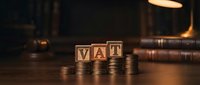 Wooden blocks spelling VAT with stacked coins on desk, representing value added tax, taxation, and financial accounting for business and accounting concepts.