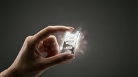 Hand holding glowing crystal or glass cube with light effects against dark background, representing innovation, technology, or digital transformation concepts.