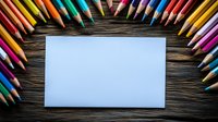 Blank white paper card surrounded by colorful wooden pencils arranged in rainbow order on dark rustic wooden table background. Creative art supplies layout.
