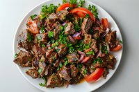 Tender braised beef chunks garnished with fresh parsley, red onions and juicy tomato slices served on white plate, appetizing meat dish for restaurant menu.