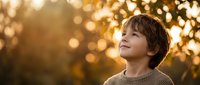 Young boy with brown hair looking up thoughtfully during golden hour sunset with warm bokeh lights creating dreamy atmosphere for childhood wonder concepts.