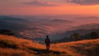 Person standing in golden grassland overlooking mountain valley at sunset with warm orange sky and rolling hills creating peaceful landscape scene.