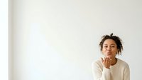 Young woman with curly brown hair and medium skin tone wearing cream ribbed sweater, blowing a kiss with hand gesture against white wall background.