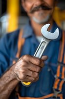 Middle-aged Hispanic mechanic in blue coveralls holding adjustable wrench, close-up view of weathered hands and tool against blurred workshop background.