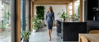 Woman in business casual attire walking through modern office space with natural light, plants, and contemporary furniture for corporate workplace design.