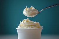 Creamy soft serve vanilla ice cream swirled on spoon above white cup against teal background, showing texture and movement in frozen dessert photography.