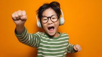 Young Asian boy with glasses and headphones celebrating with raised fist against vibrant orange background, expressing joy and excitement in striped green shirt.