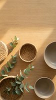 Natural wicker baskets and ceramic bowls arranged with fresh eucalyptus branches on warm wood surface with soft sunlight shadows for home decor styling.