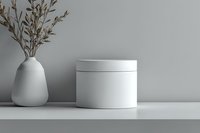 Minimalist white cosmetic jar mockup with dried plant in ceramic vase against gray wall, perfect for skincare product presentation.