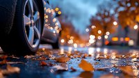 Car wheel on wet asphalt with fallen autumn leaves and blurred bokeh lights creating magical evening atmosphere, low angle view at street level.