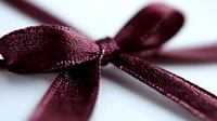 Elegant burgundy satin ribbon bow with delicate texture against white background, macro close-up view showing fine fabric details and soft shadows.