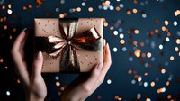Hands holding wrapped present with metallic bow against dark background with glowing orange and blue bokeh lights creating magical holiday atmosphere.
