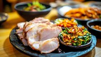 Succulent sliced pork belly with crispy skin served alongside colorful stir-fried vegetables on black stone plate, showcasing Asian fusion cuisine preparation.